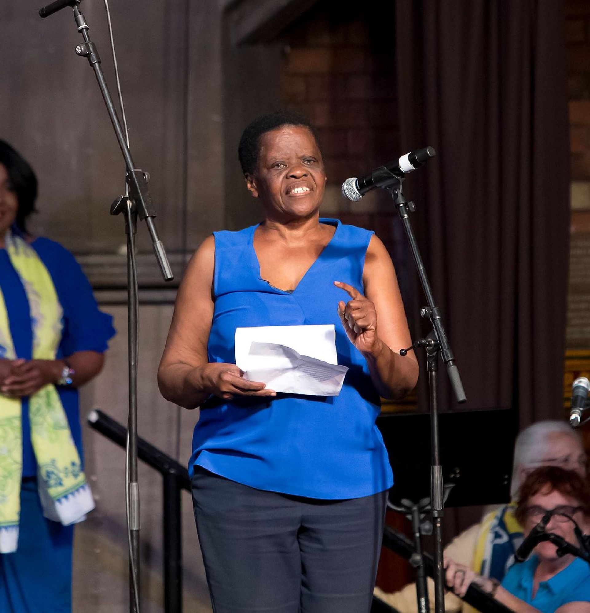 A woman in a blue shirt stands in front of a microphone holding a piece of paper