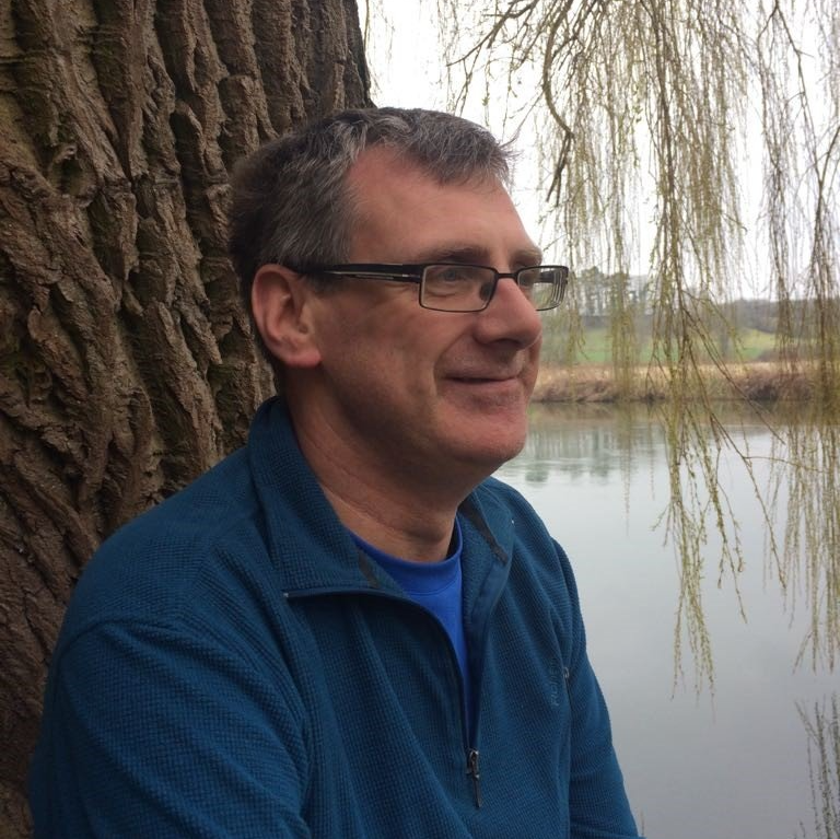 A man wearing glasses is leaning against a tree near a body of water