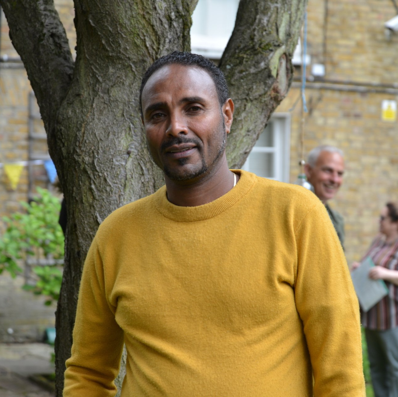 A man in a yellow sweater stands in front of a tree