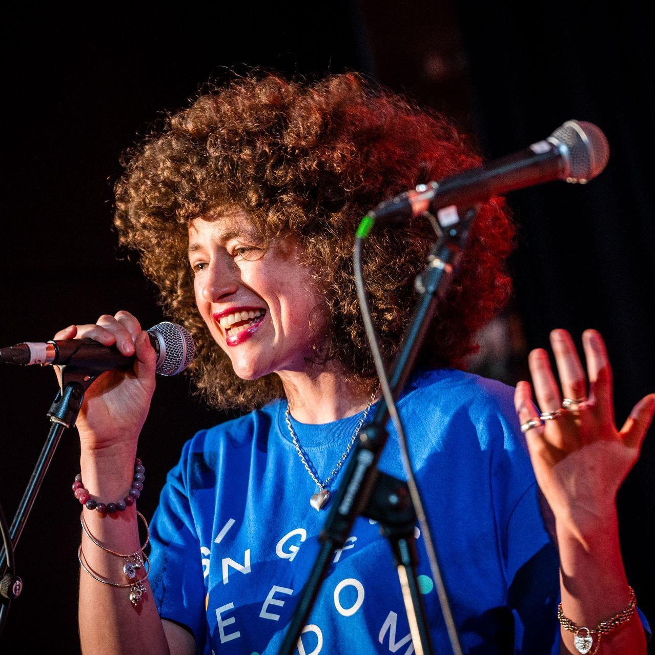 A woman with curly hear, wearing a blue t-shirt, is smiling and speaking into a microphone