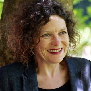 A woman with curly hair is smiling while leaning against a tree.