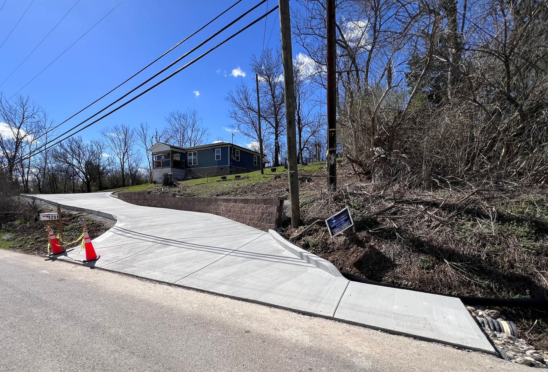 New concrete driveway rising beside a road, with orange cones and power lines on a sunny hillside