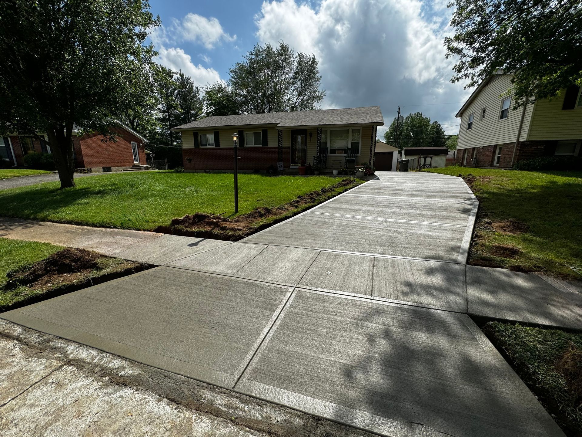 Concrete driveway leading to a single-story brick house with a small front lawn and trees.