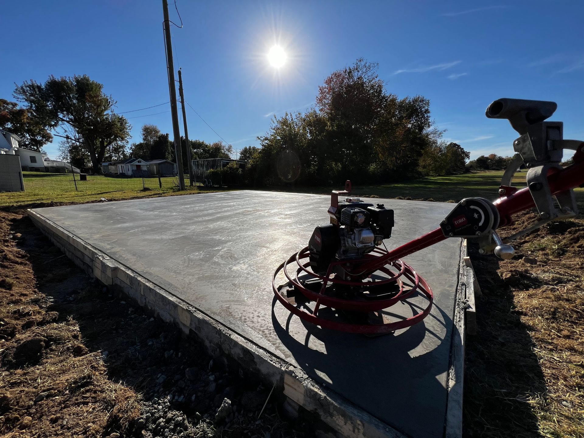 A worker uses a concrete float to smooth wet cement being poured from a truck, with a yellow loader in the background.