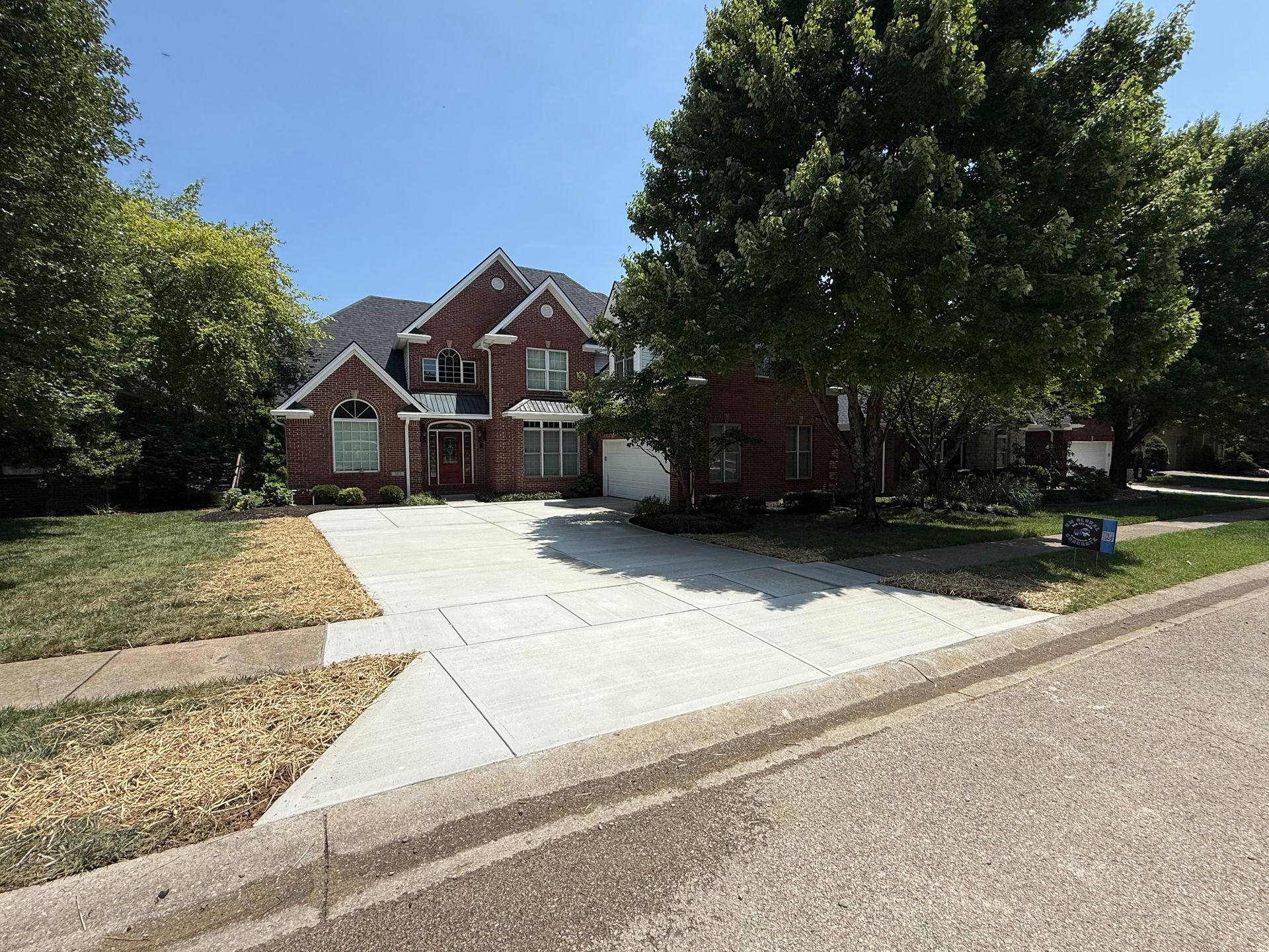 Brick suburban house with a white driveway, large trees, and a clear blue sky