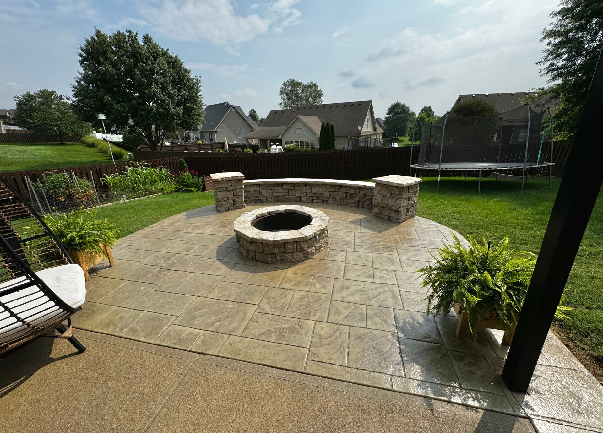 Backyard patio with a circular stone fire pit, lawn, plants, and suburban houses in the background