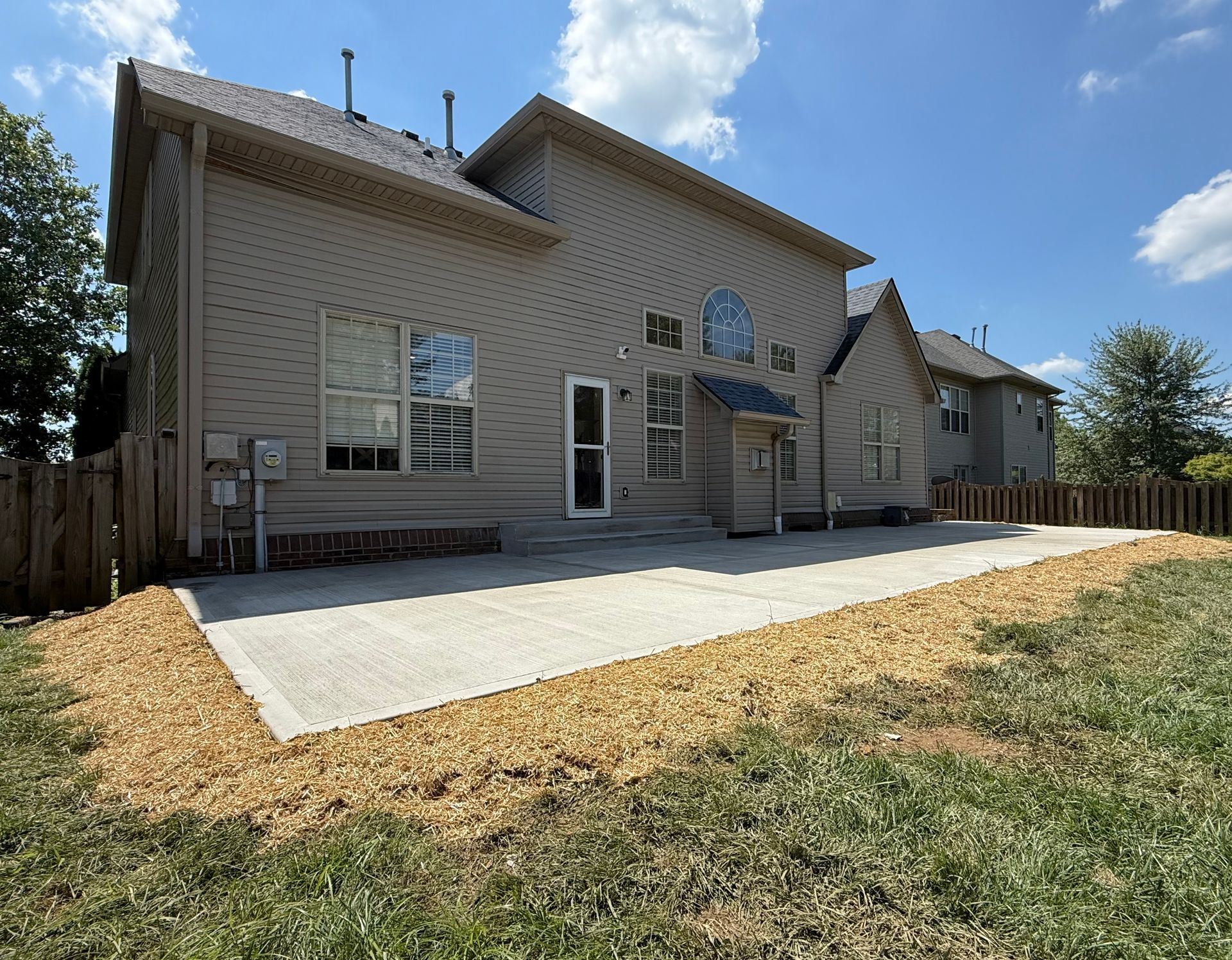 Rear view of a beige house with a concrete patio and fenced backyard under a partly cloudy sky