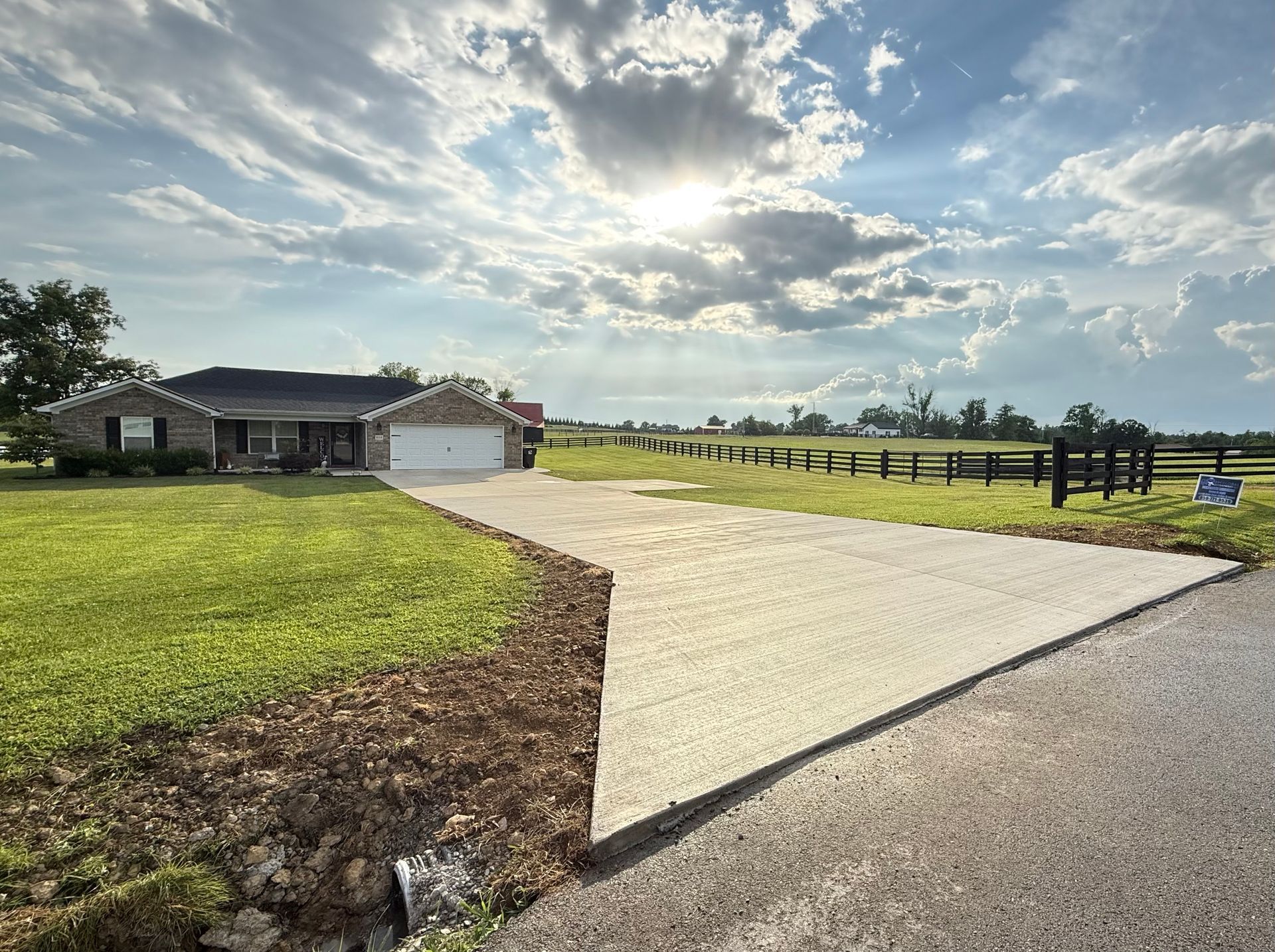 Country driveway leading to a house and garage beside a green field under a cloudy sky