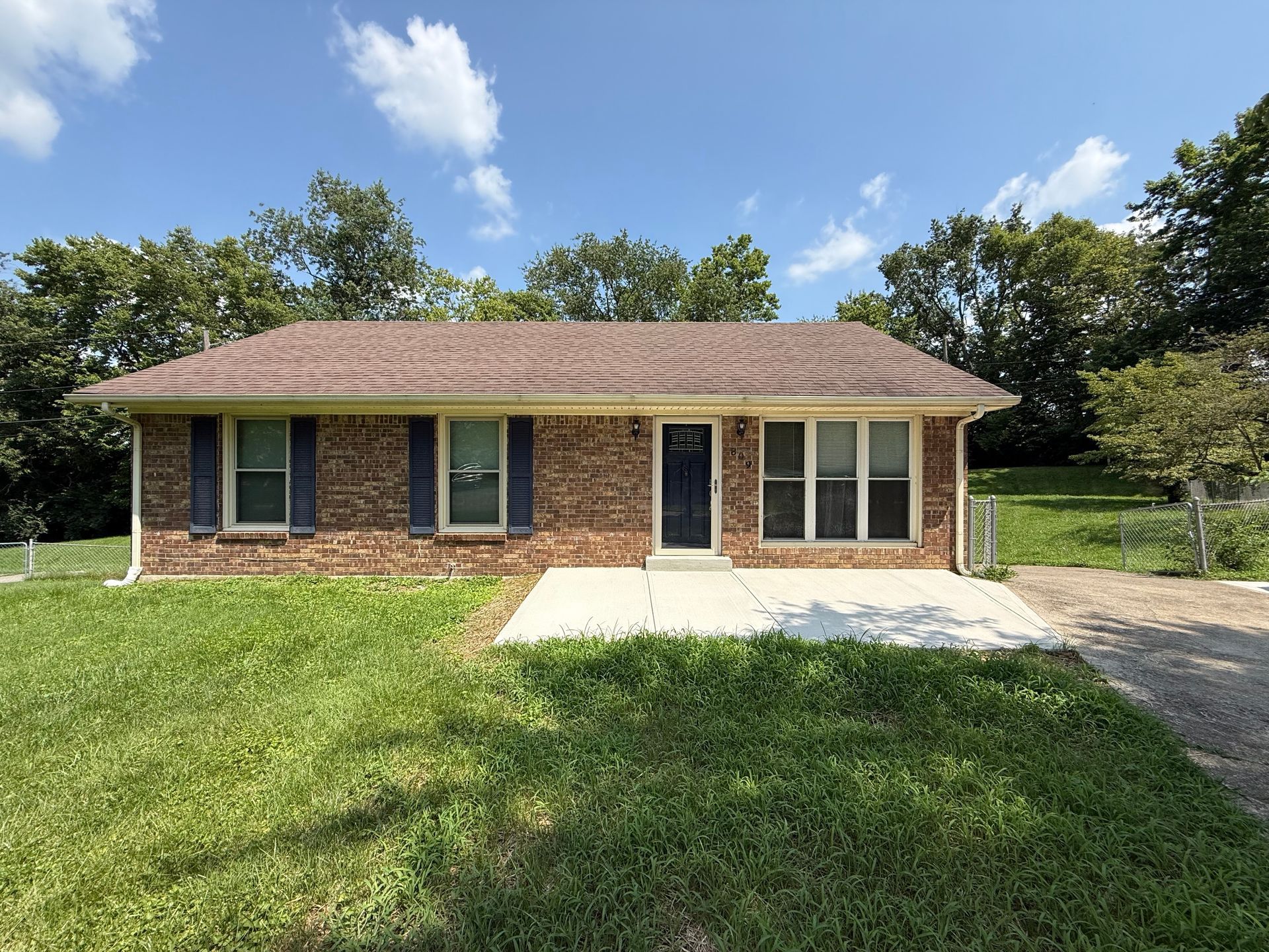 Single-story brick house with a brown roof, front lawn, and concrete patio under a blue sky