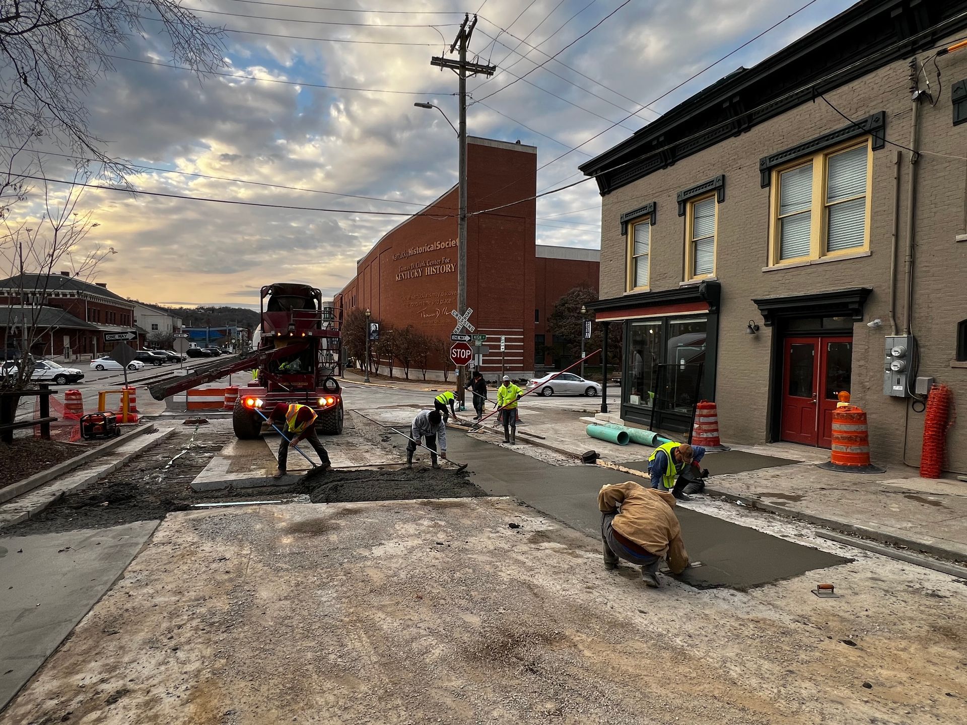 Street construction scene with workers, trucks, and barricades on a torn-up road beside buildings.