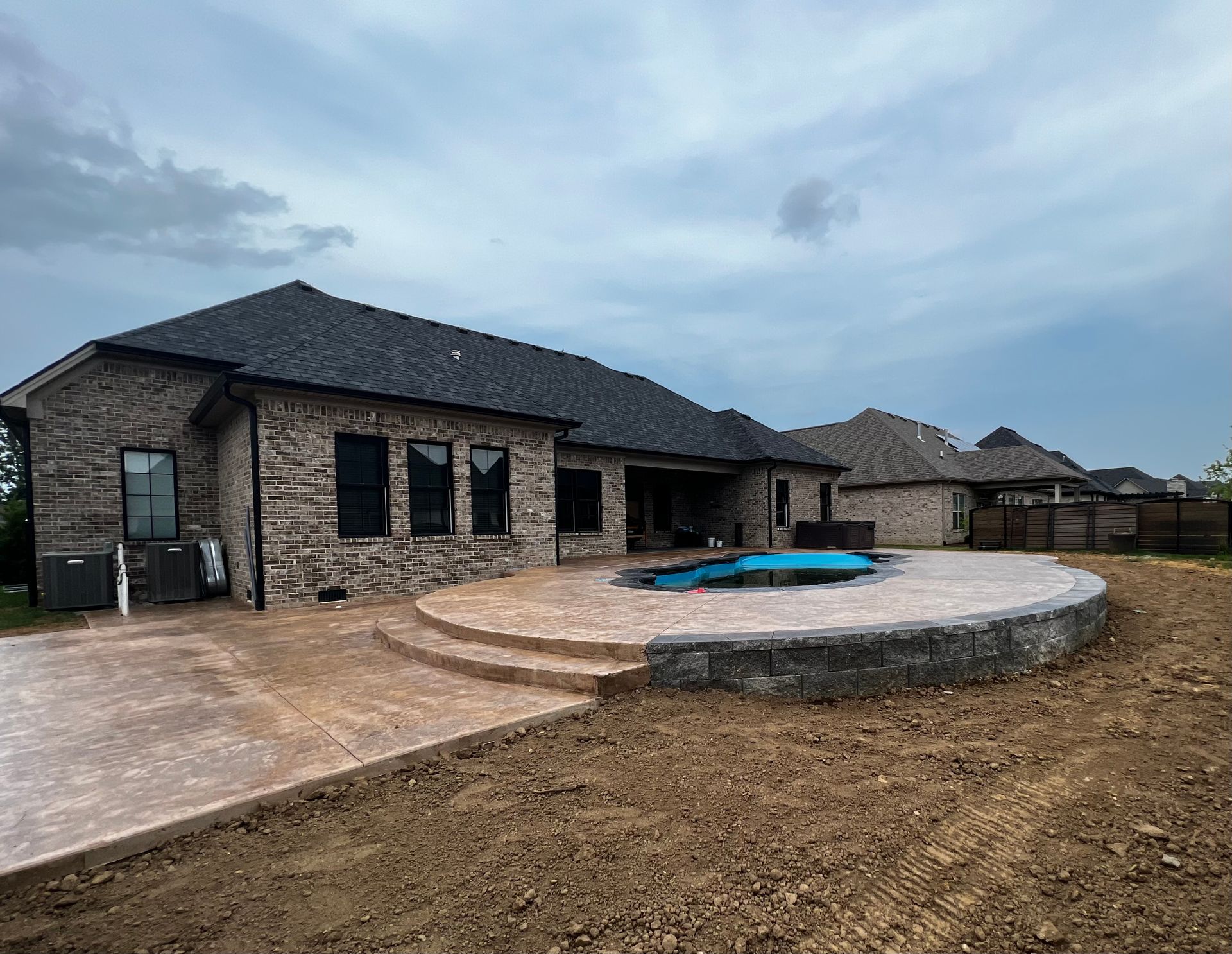 Stone house with dark roof, concrete patio, and a small round blue pool in a bare yard under cloudy sky