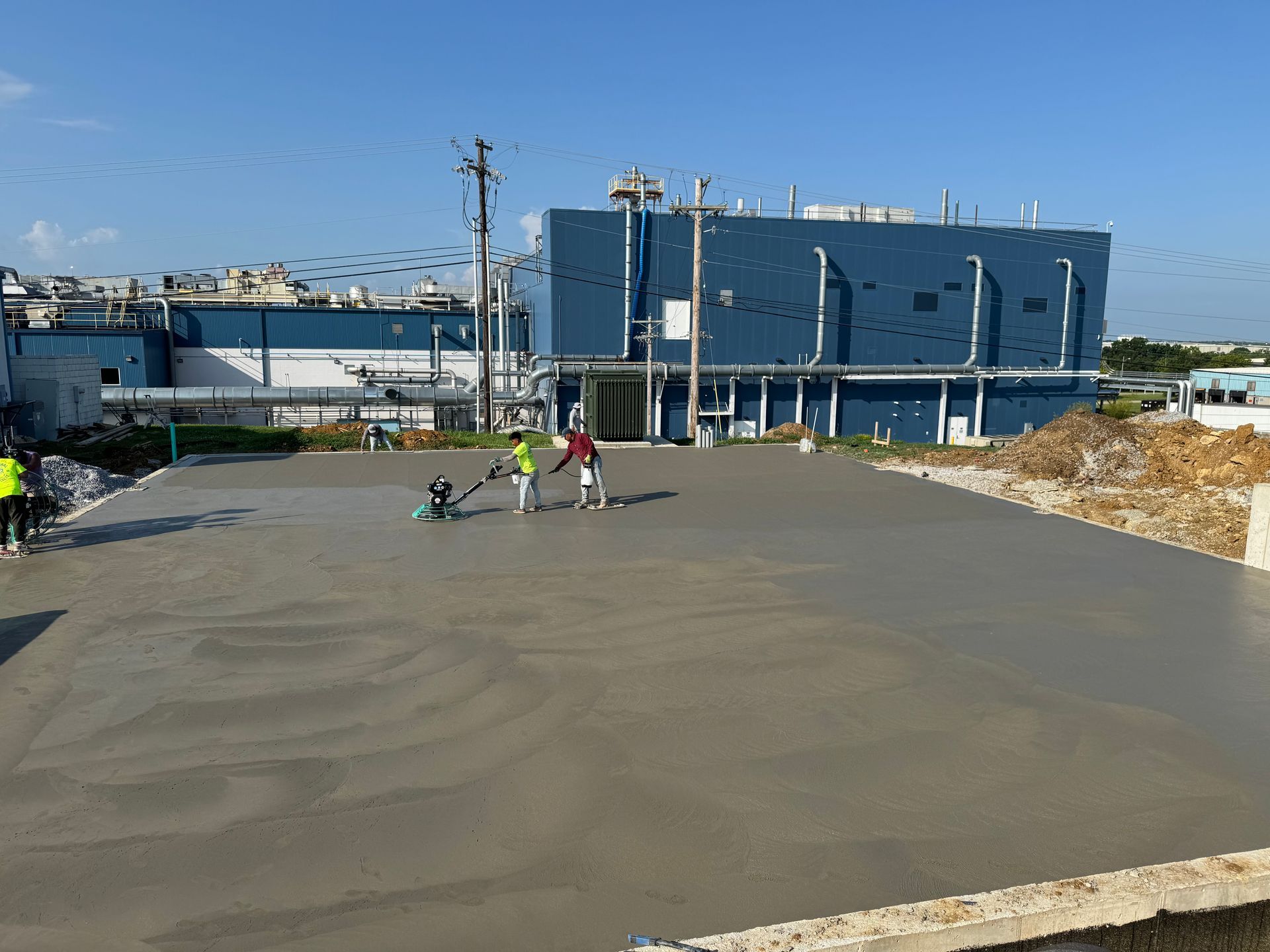 People standing on a large concrete lot near blue industrial buildings under a clear sky