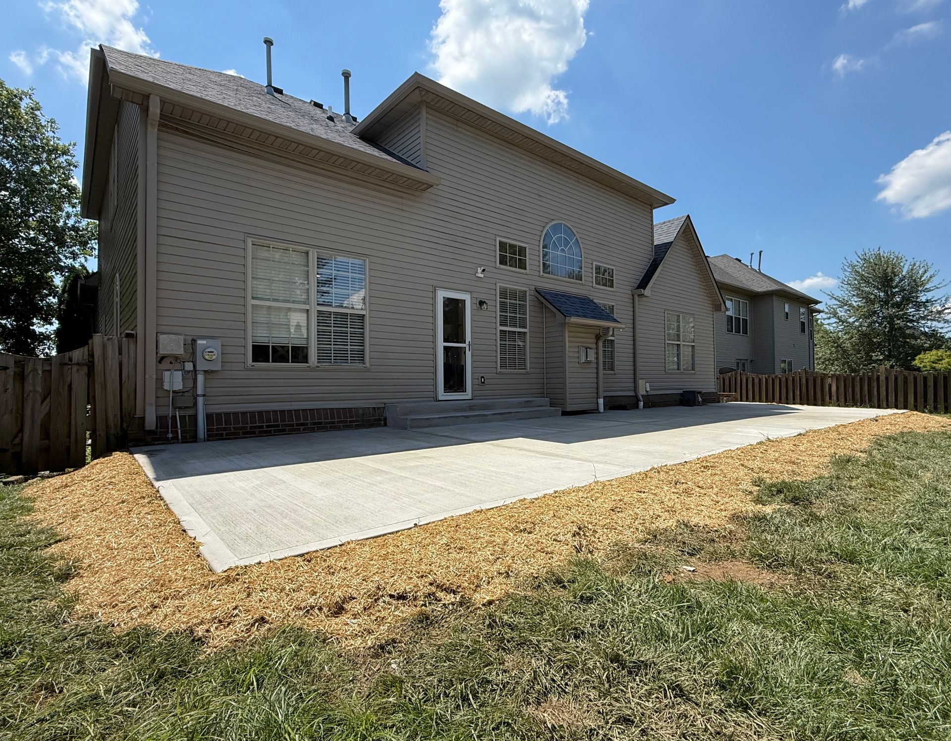 Backyard patio beside a beige house with a concrete slab and fenced yard under a blue sky