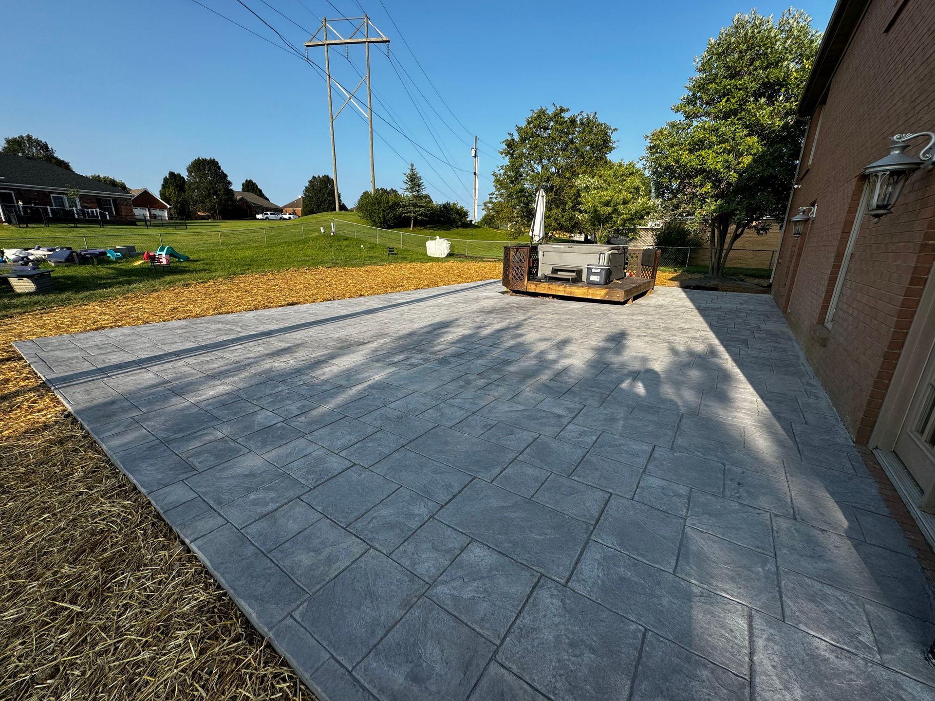 Paver patio under construction beside a brick house, with a compacting machine and piles of sand.