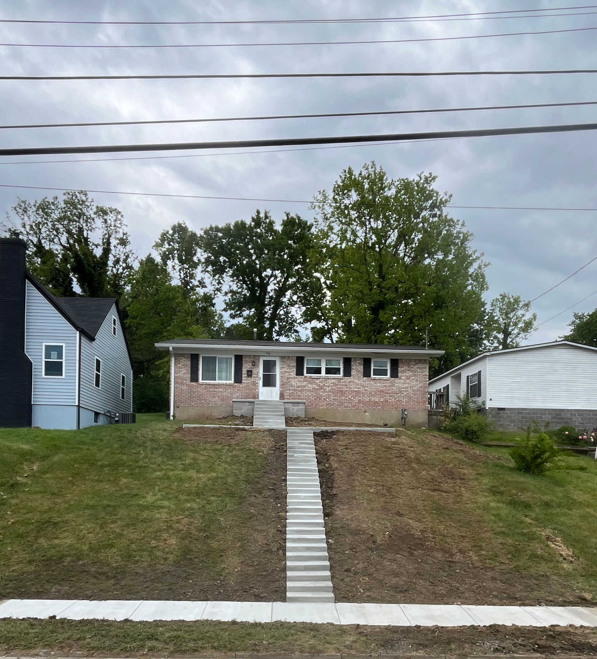 Small brick house on a grassy hill with a concrete stair path, flanked by neighboring homes and overhead power lines.