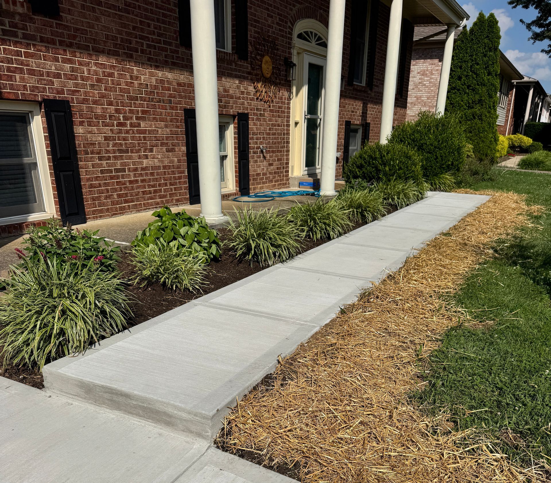 Sidewalk beside a brick building with white columns, bordered by mulch and green shrubs