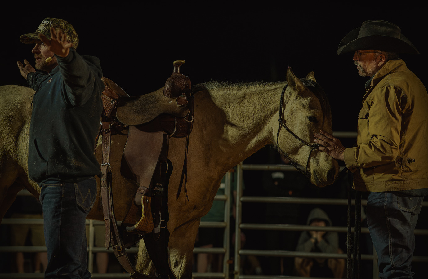 Cowboy standing beside saddled horse in a dimly lit rodeo pen at night