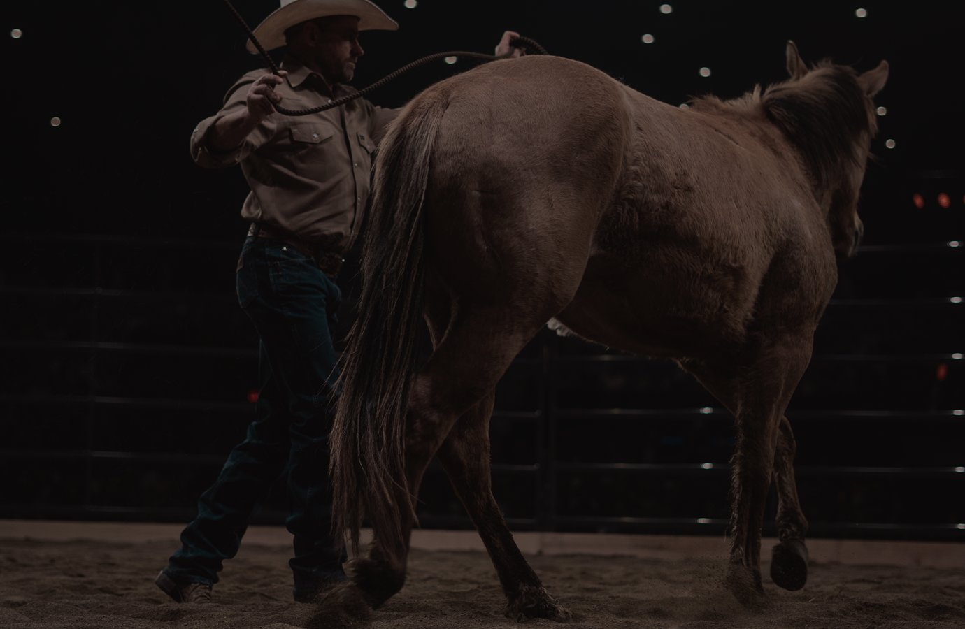 Cowboy in a hat standing beside a saddled horse in a dimly lit arena