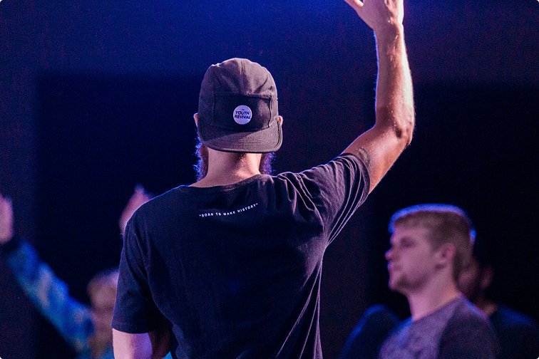 Person on stage with arm raised, wearing a cap under blue-purple concert lighting.
