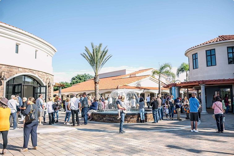Crowded outdoor plaza with people gathering near shops under a clear blue sky
