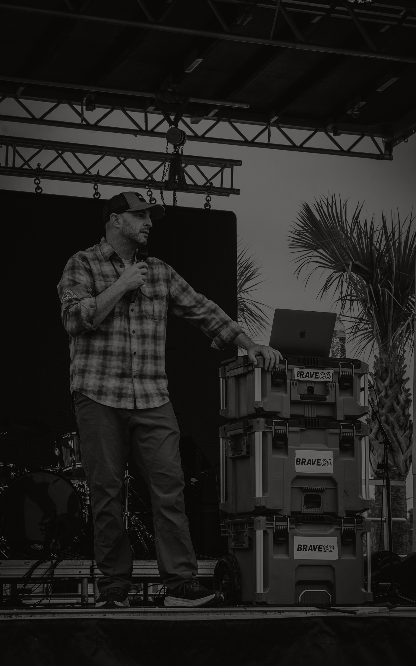 Man speaking on an outdoor stage beside audio equipment, holding a microphone under a truss roof.
