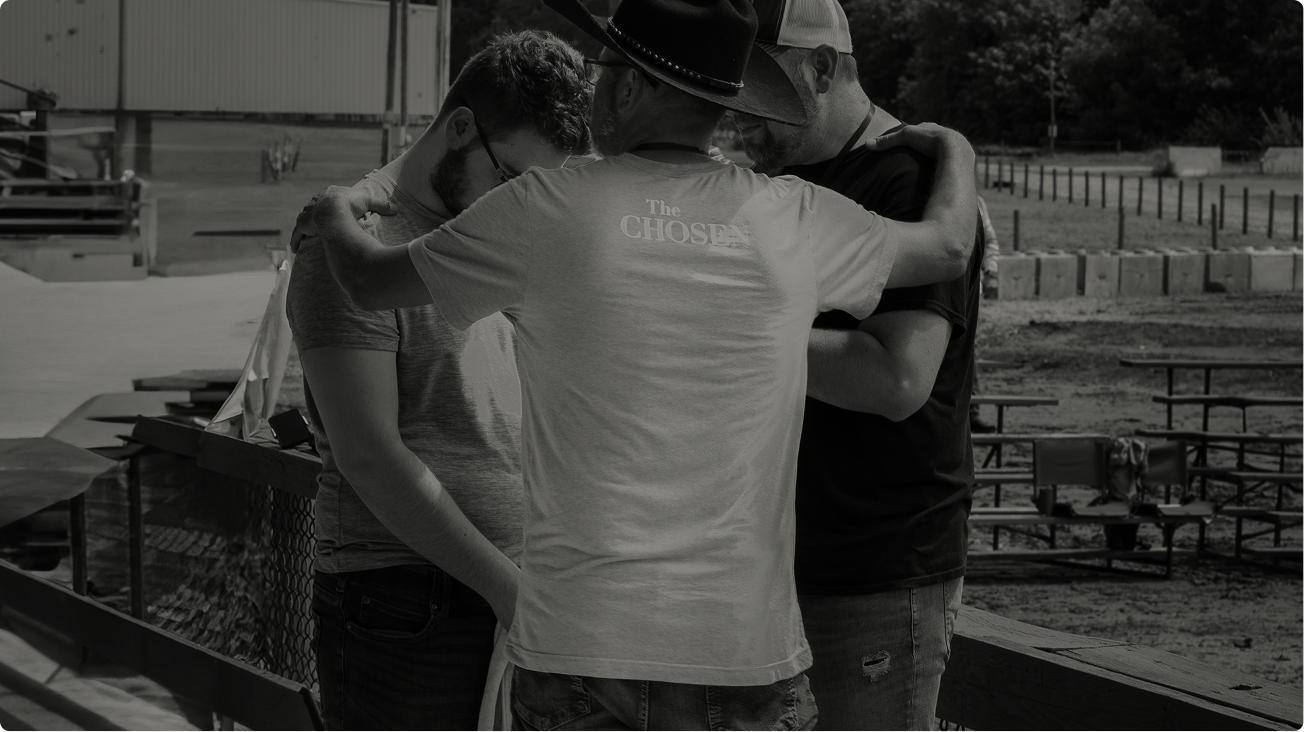 Three people stand close together outdoors, one with an arm around another near a fenced area.