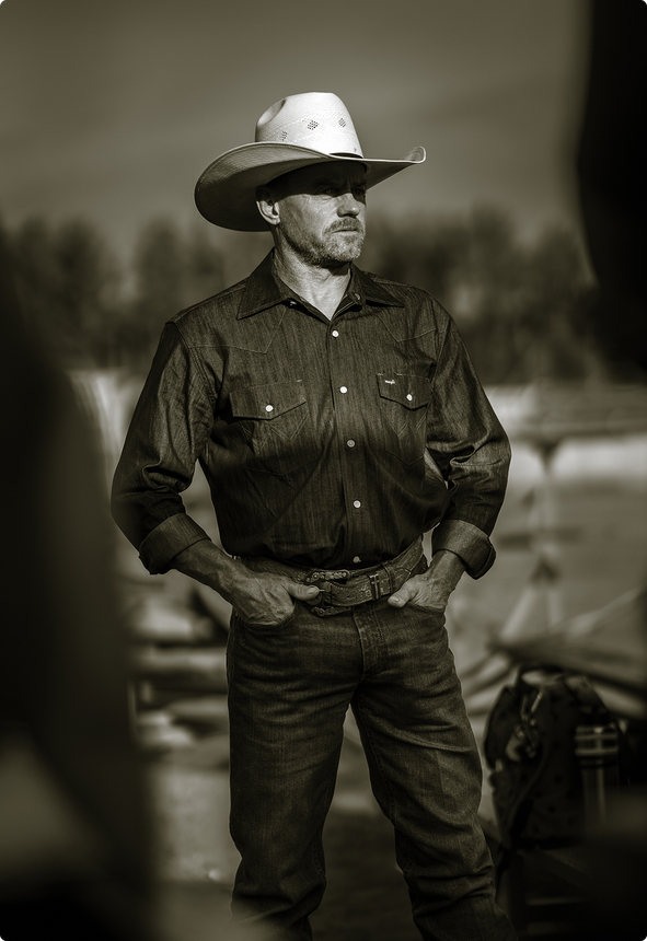 Man in a cowboy hat standing outdoors with hands in pockets, sepia-toned.