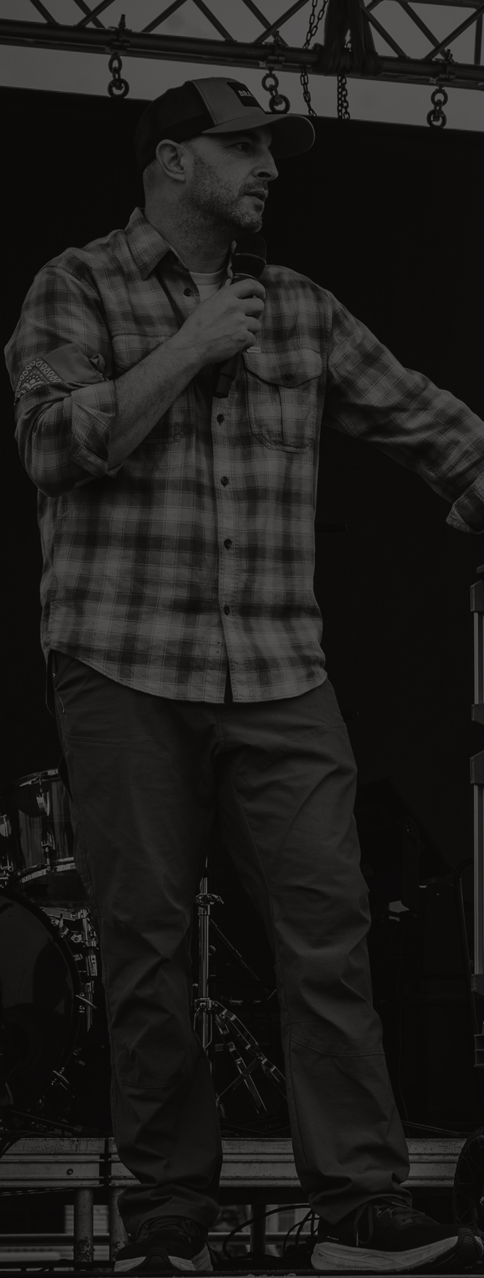 Man speaking on an outdoor stage beside audio equipment, holding a microphone under a truss roof.