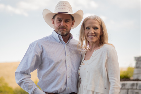 Two people standing outdoors, the man in a white cowboy hat and the woman smiling beside him.