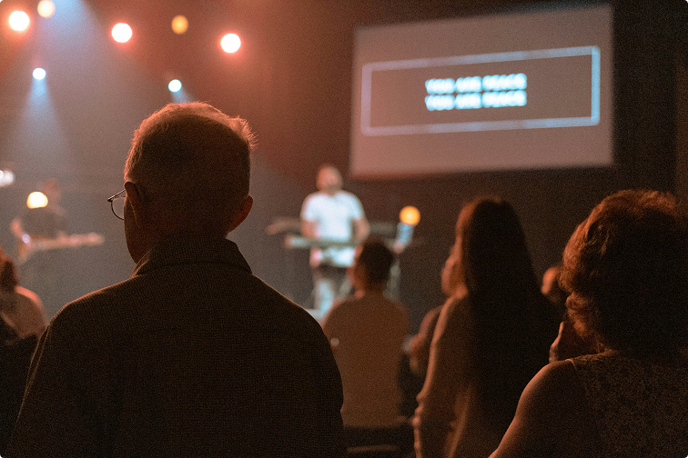 Audience watching a speaker on a dimly lit stage with a projected screen in the background