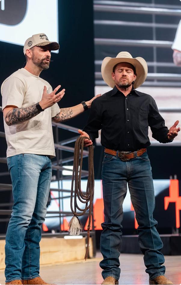 Two men standing on a stage, one in a white T-shirt and cap, the other in a cowboy hat and black shirt.
