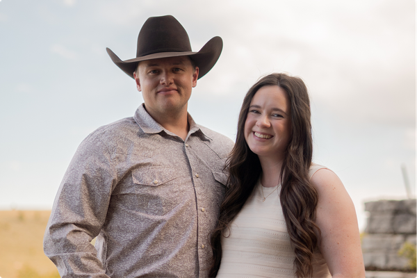 Couple smiling outdoors, with a man in a cowboy hat and a woman in a white dress.