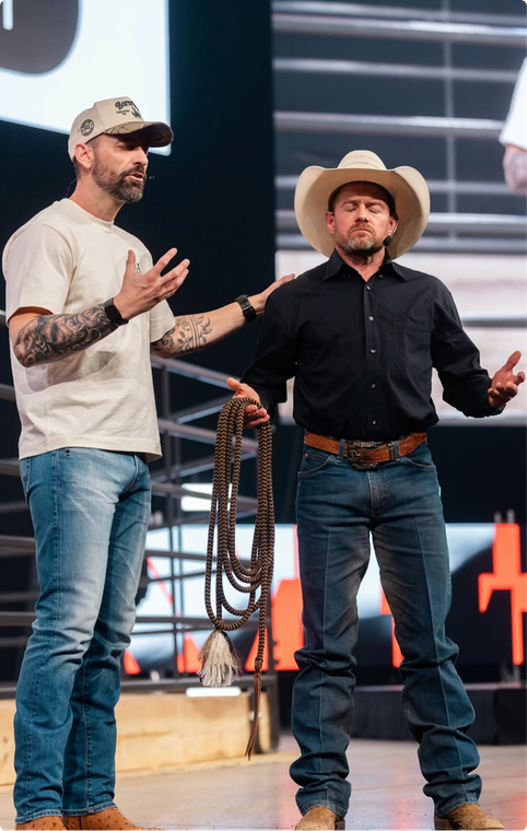 Two men standing on a stage, one in a white T-shirt and cap, the other in a cowboy hat and black shirt.