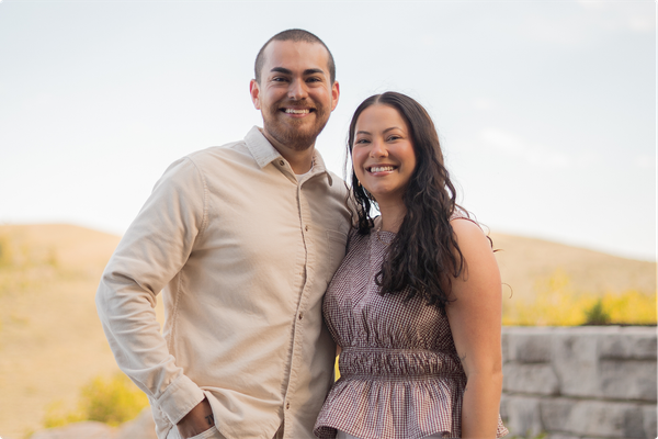 Smiling couple standing outdoors near a stone wall, with a bright sky and hills behind them.