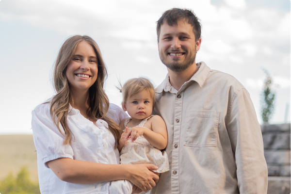 Family of three smiling outdoors, with a baby held between two adults against a bright sky.