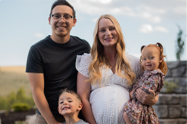 Family of four outdoors, smiling with a pregnant woman holding a toddler at sunset