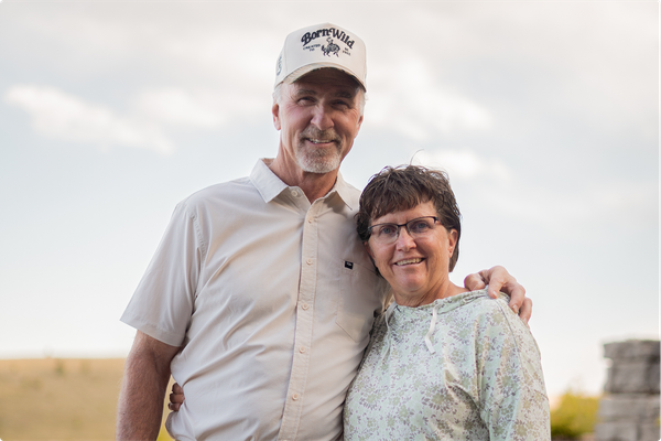 Smiling couple standing outdoors in a sunny field, with the man’s arm around the woman