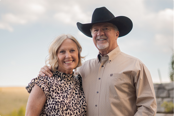Smiling couple in cowboy hats standing together in a grassy field at sunset