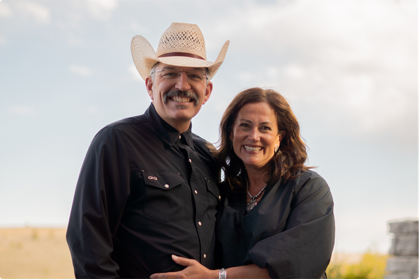 Smiling couple standing outdoors at sunset, the man in a white cowboy hat and black shirt hugging the woman.