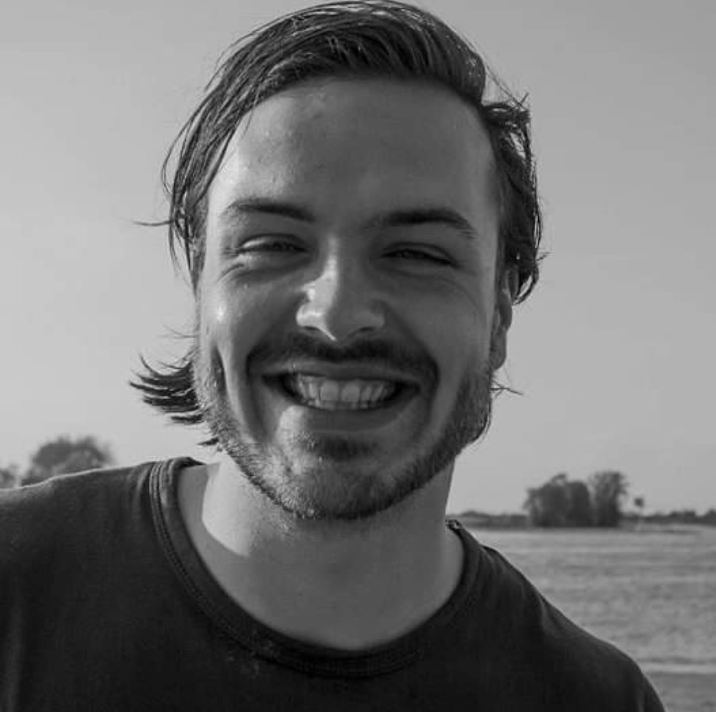 Lachende man met donker haar, kuiltjes in zijn wangen en een baard, buiten met een heldere lucht op de achtergrond.