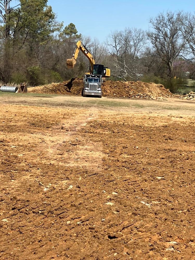 A yellow excavator digs into a large dirt mound behind a silver truck in a sunny, rural construction area.