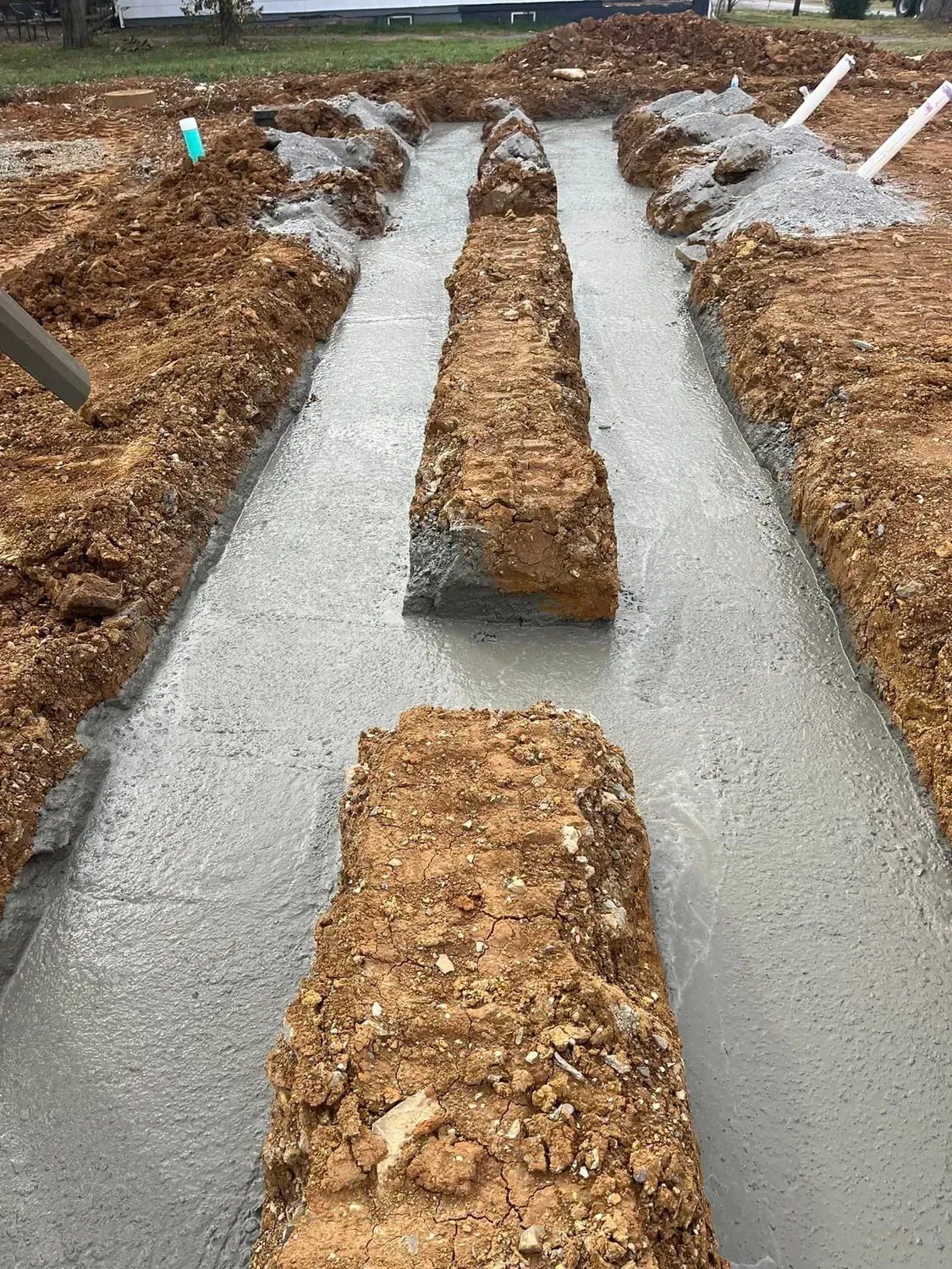 Freshly poured concrete footings in trenches with mounds of dirt between them at a construction site.