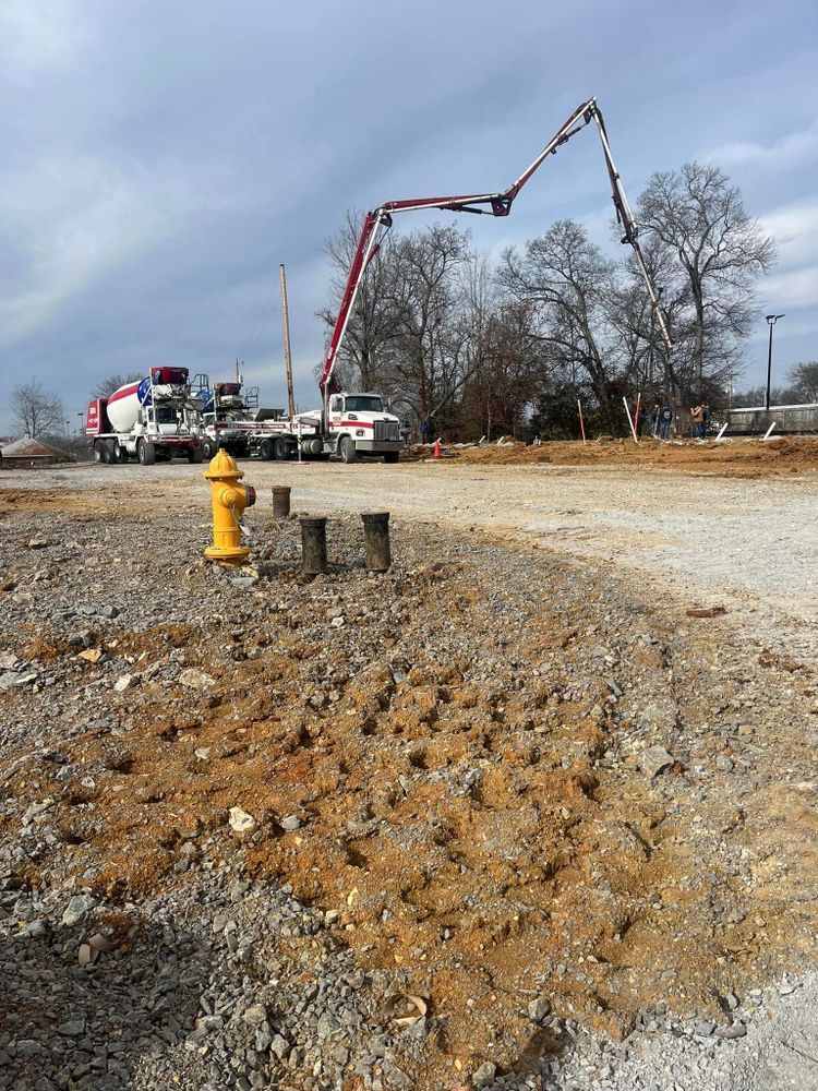 A concrete pump truck with its boom extended stands on a gravel lot near a yellow fire hydrant under a cloudy sky.