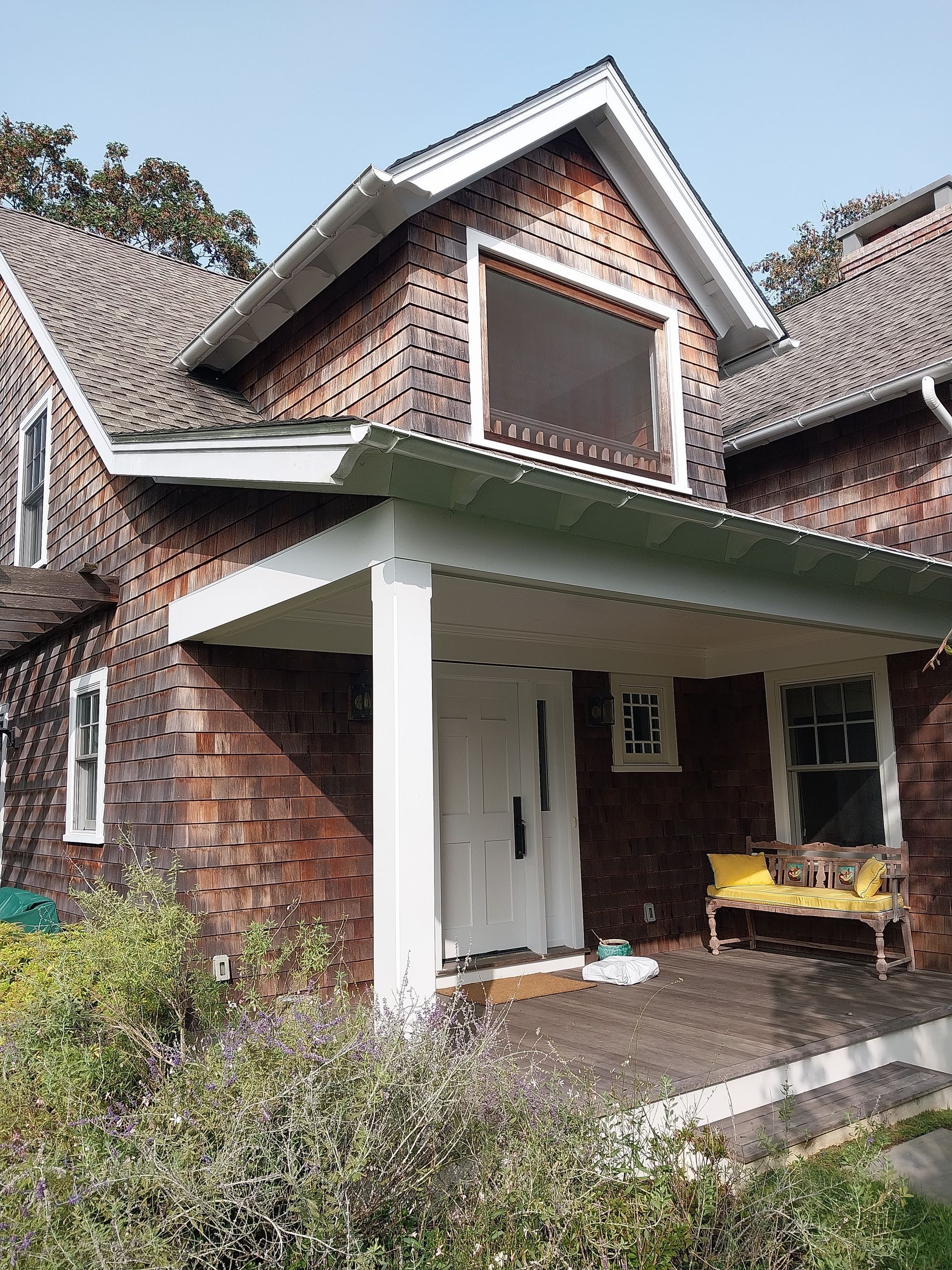 Two-story house with brown shingles, a white porch, and a yellow bench. The house has white trim and small windows.