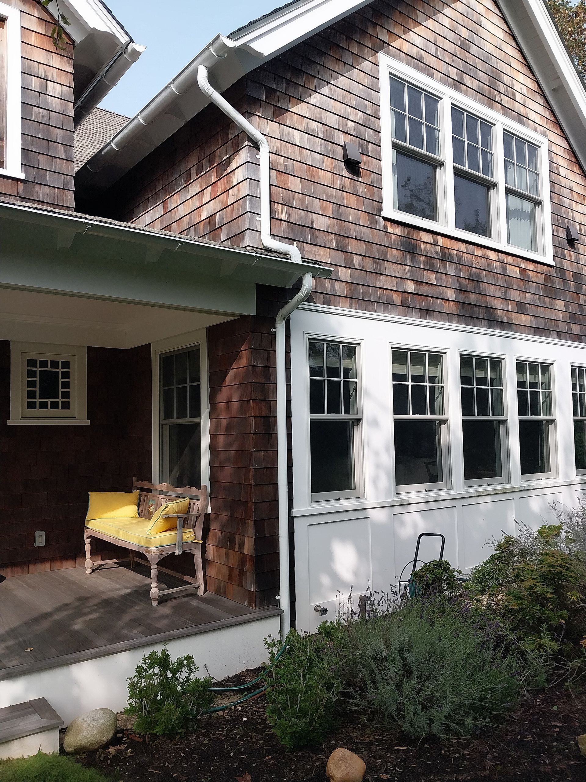 Shingled house with a porch and multiple windows. A yellow bench sits on the porch, and plants are in the foreground.