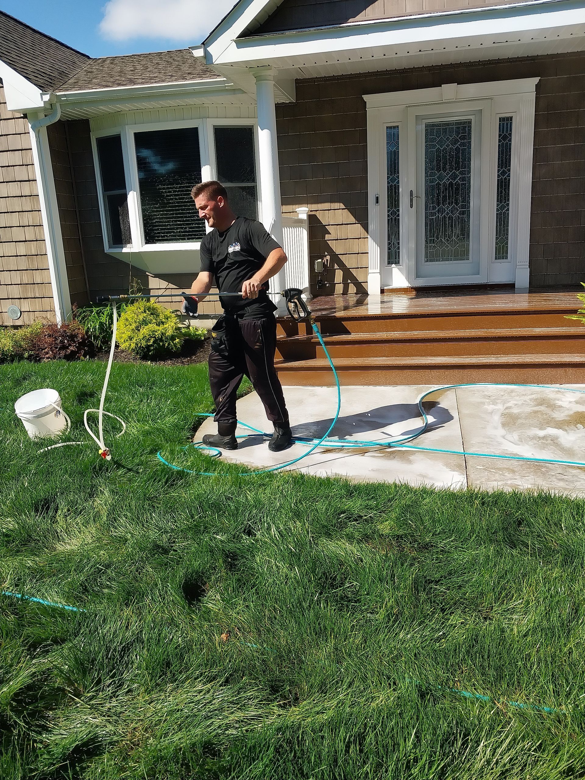 A man in black clothing is power washing a concrete walkway in front of a house with brown siding.