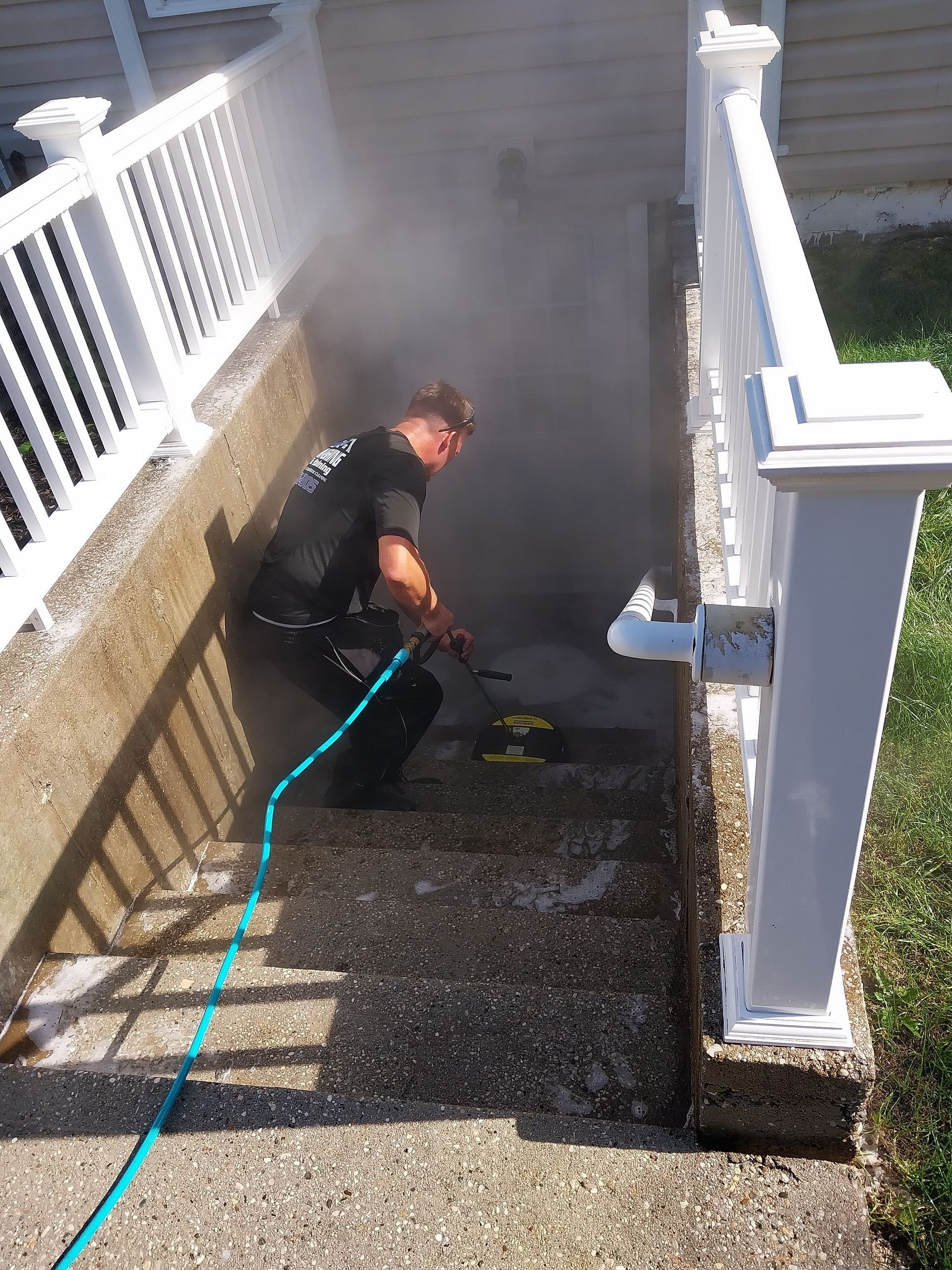 A person pressure washes concrete stairs, creating a cloud of mist. The stairs have white railings.