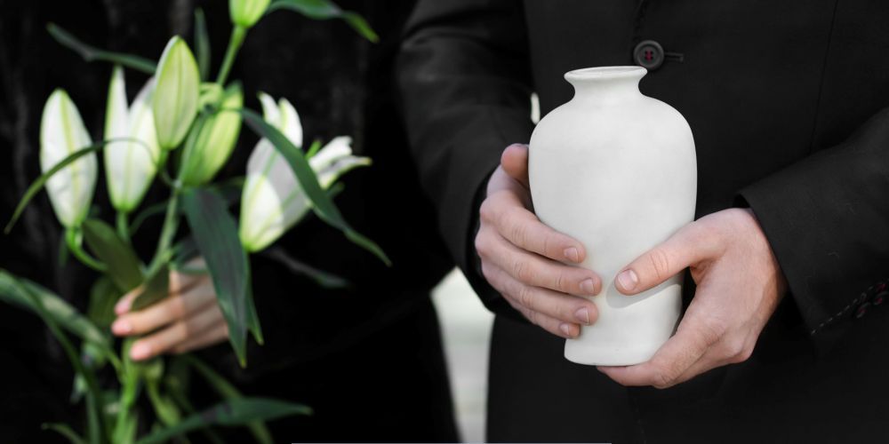 Person holding a white urn beside white lilies at a memorial service for Compassionate Cremation Services in Marlton, NJ.