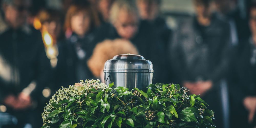 Metal cremation urn displayed on a floral arrangement with mourners in the background during Compassionate Cremation Services in Marlton, NJ.