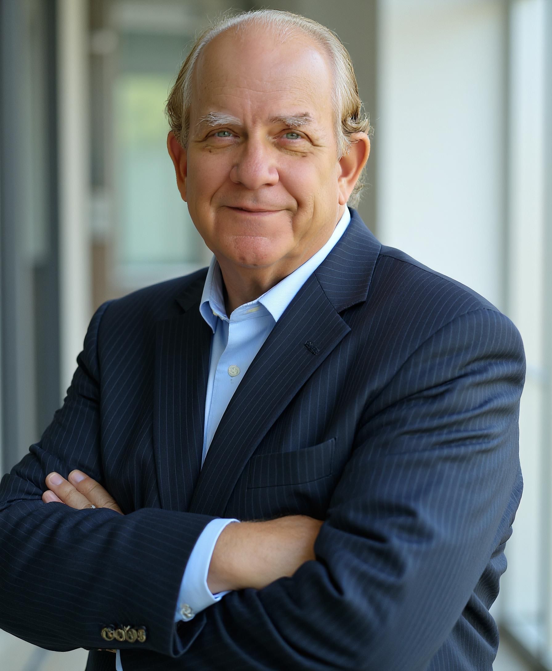 Portrait of a man in a dark suit with arms crossed, standing indoors and smiling gently.